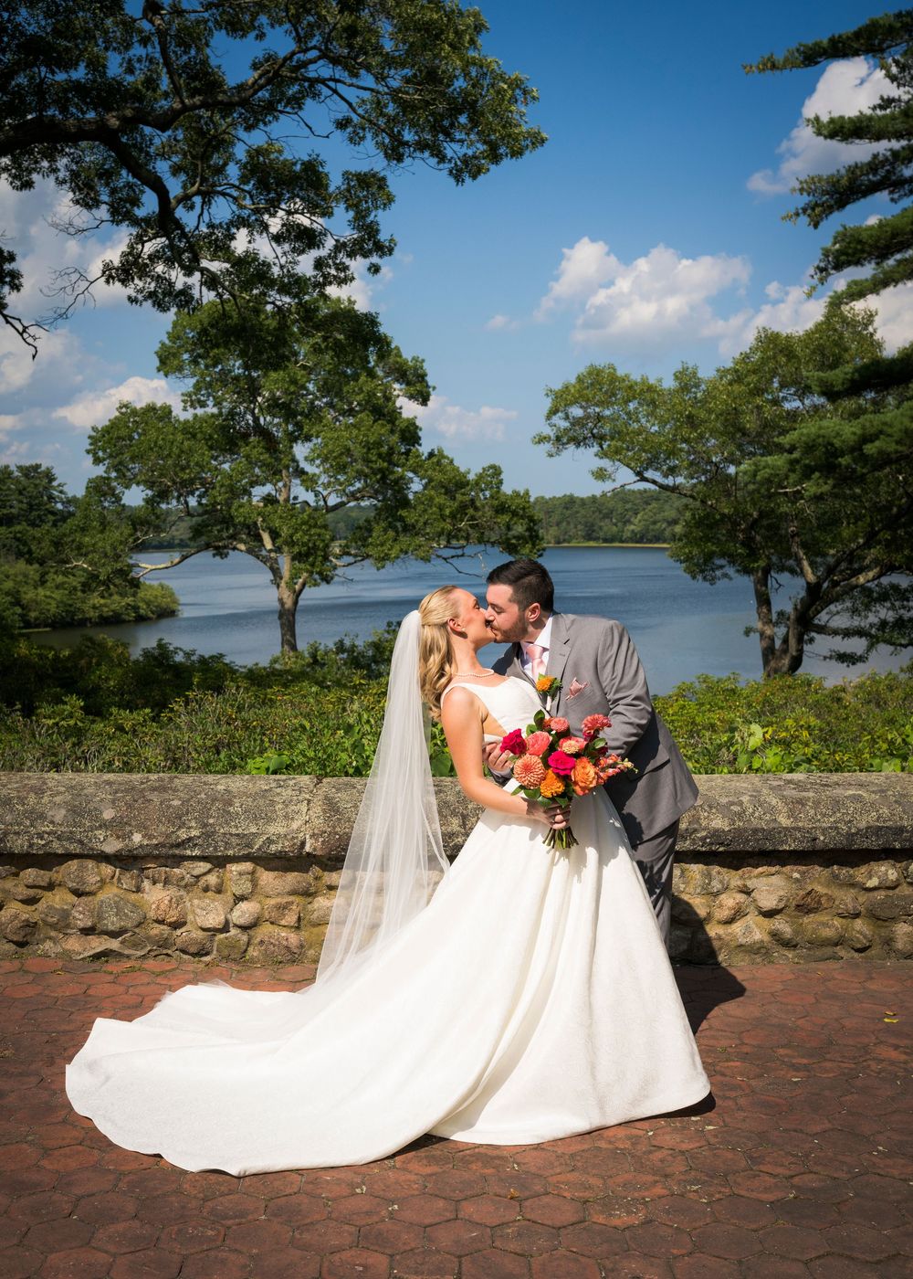 Bride and groom share a kiss on a brick terrace with a stunning lake and blue sky backdrop.