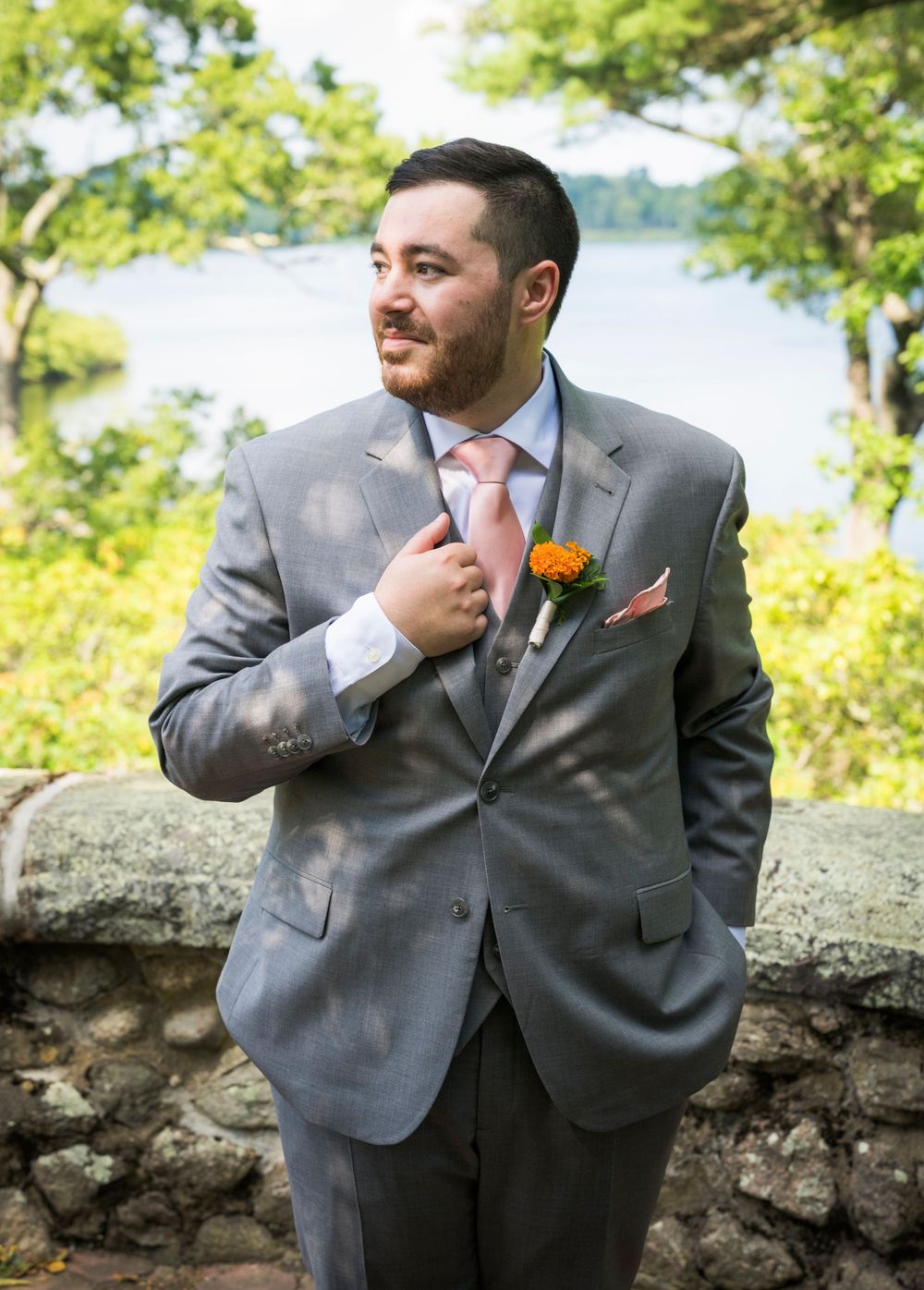 Dapper groom in a gray suit with pink tie and orange boutonniere adjusts his jacket near a stone wall.