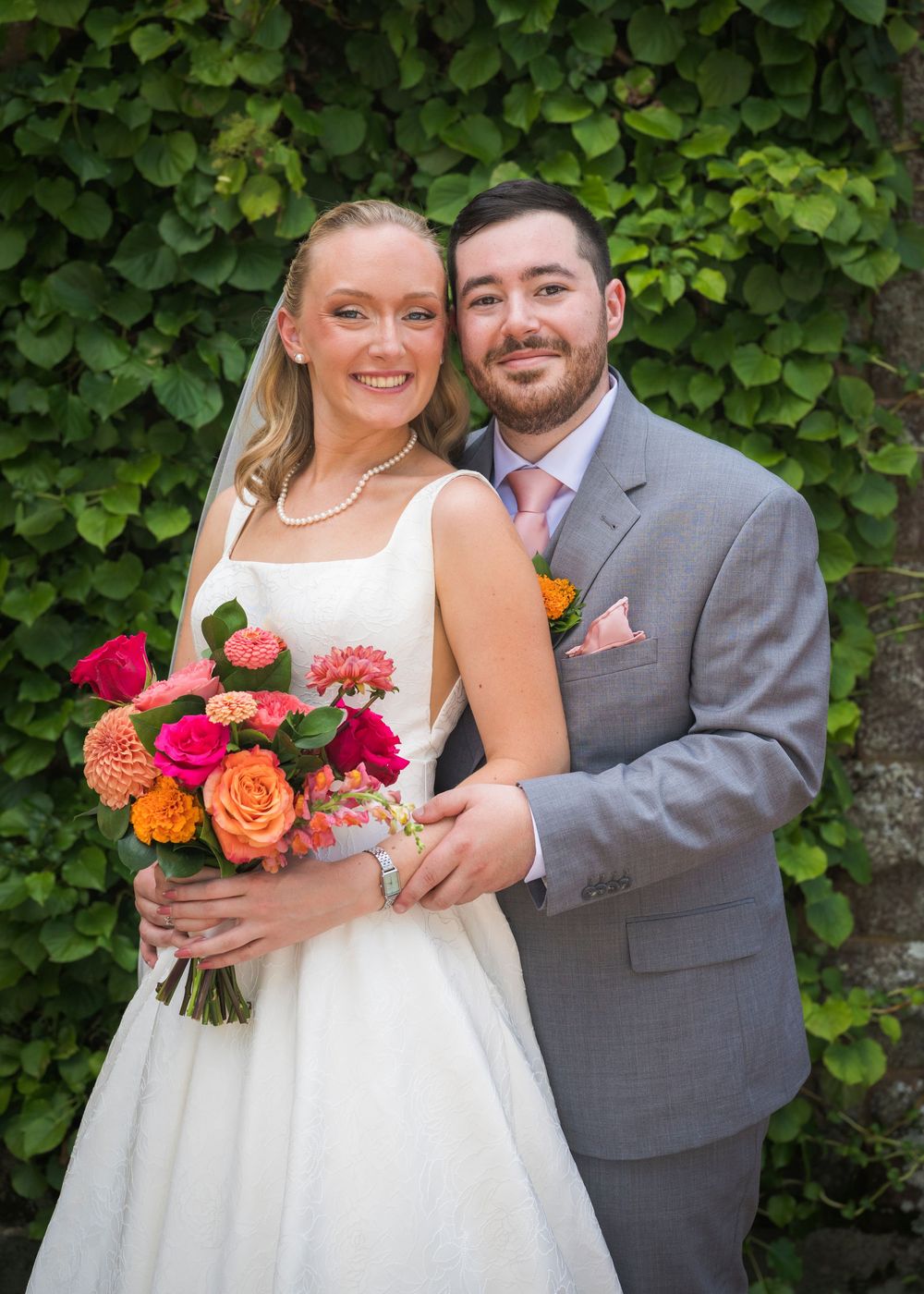 Bride and groom posing together outdoors, bride holding vibrant pink and orange floral bouquet, groom in gray suit with pink tie.