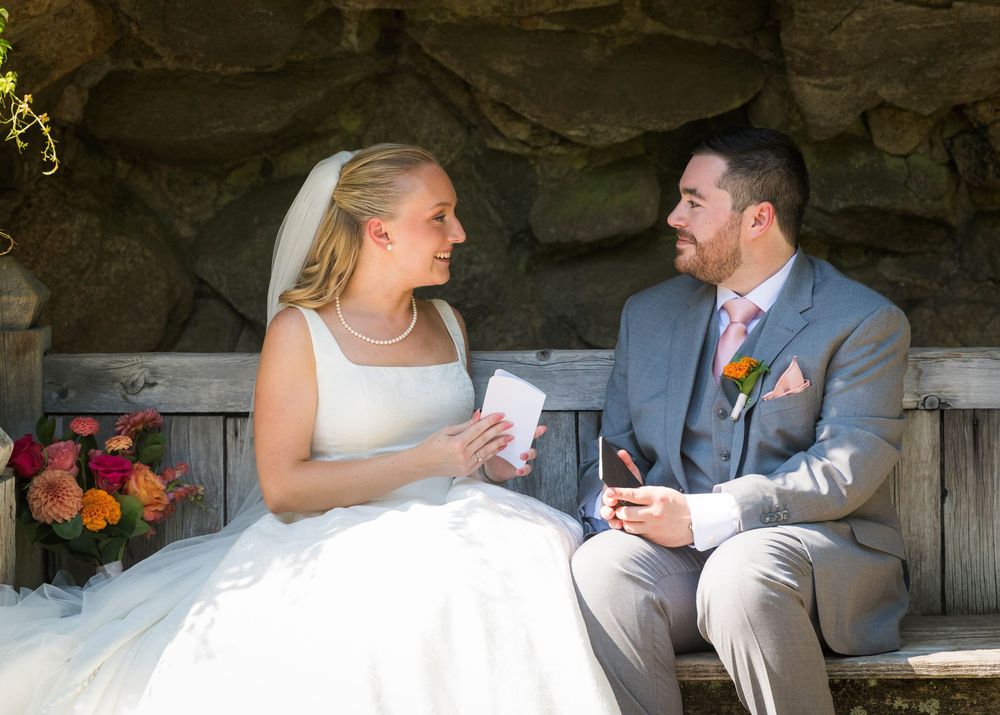 Bride and groom exchange vows on a bench, bride holding note cards, surrounded by colorful floral arrangements.
