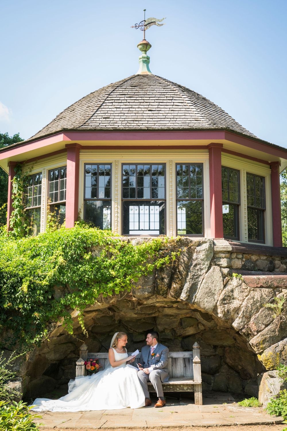 Bride and groom sit on a bench beneath a charming octagonal gazebo surrounded by lush greenery.