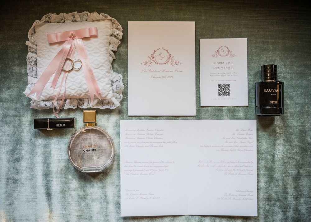 Wedding flat lay featuring invitation suite, ring pillow, and perfume bottles on elegant gray surface.