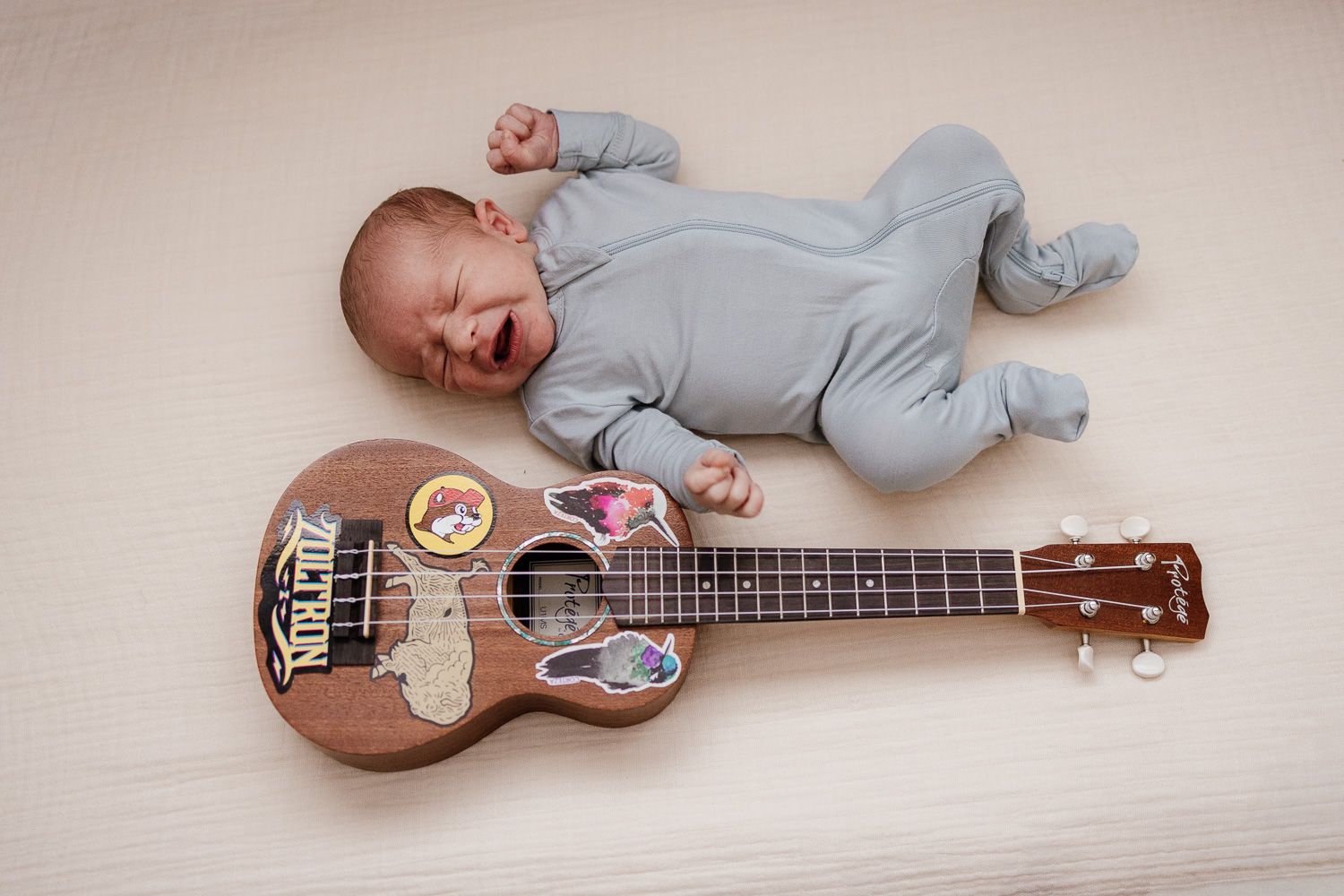 Crying newborn baby looks like a tiny rockstar while lying next to a ukulele with stickers on a white background.