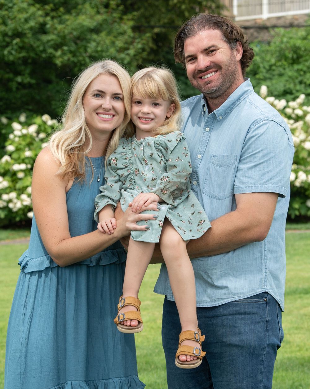 A smiling family of three poses outdoors, with parents in blue outfits holding a young toddler girl in a floral dress.