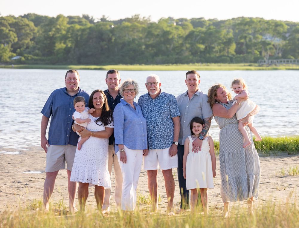 Family group portrait on sandy beach by water, smiling adults and children in coordinated blue and white outfits.