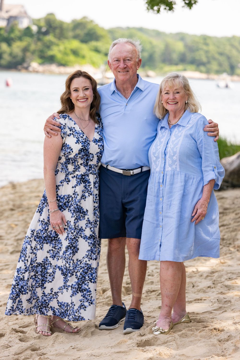 Three smiling people pose together on a sandy beach, wearing blue outfits, with water and trees in the background.