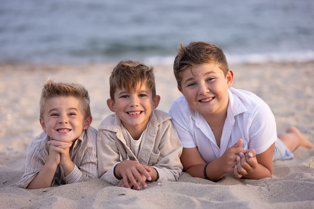 Three smiling boys lying on sandy beach, posing together for a cheerful seaside family photo.