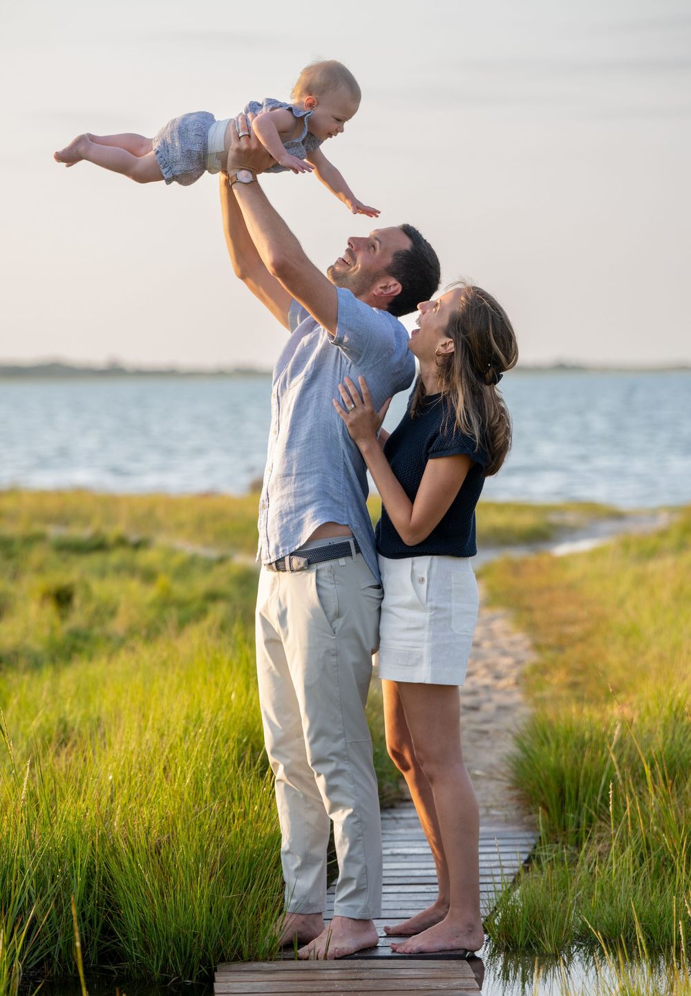 A joyful couple stands on a wooden dock by the water, lifting their baby in the air on a sunny day.