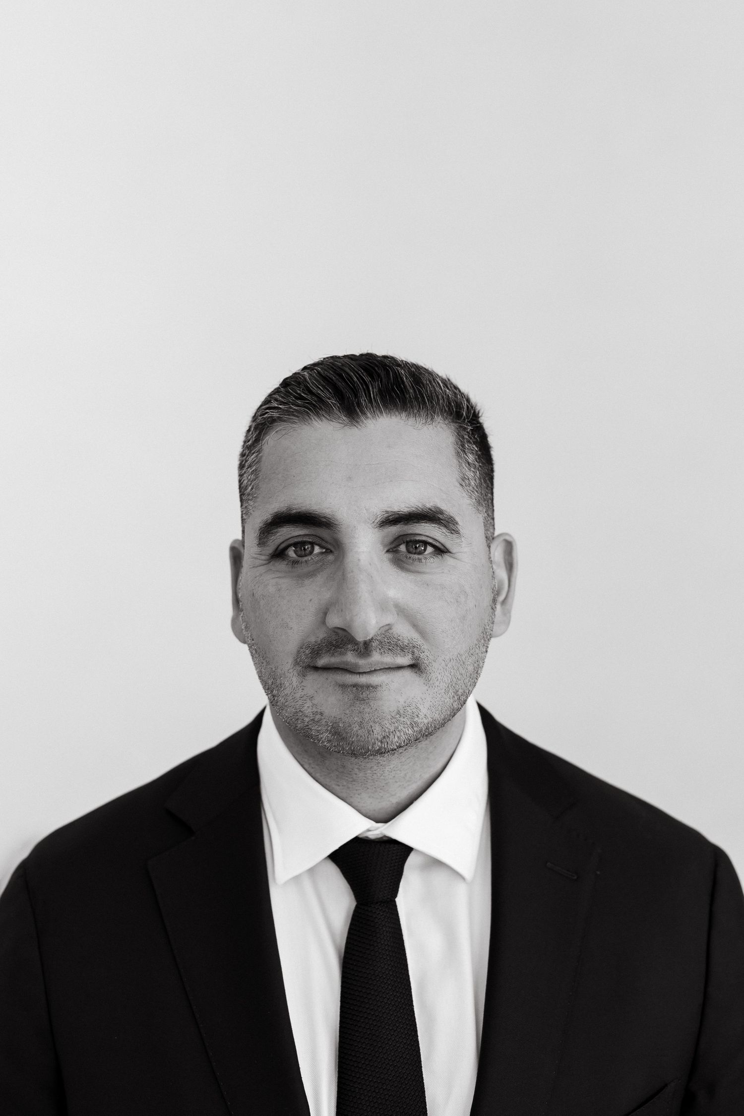 Black and white professional headshot of a man in a suit and tie with short dark hair and a subtle smile.
