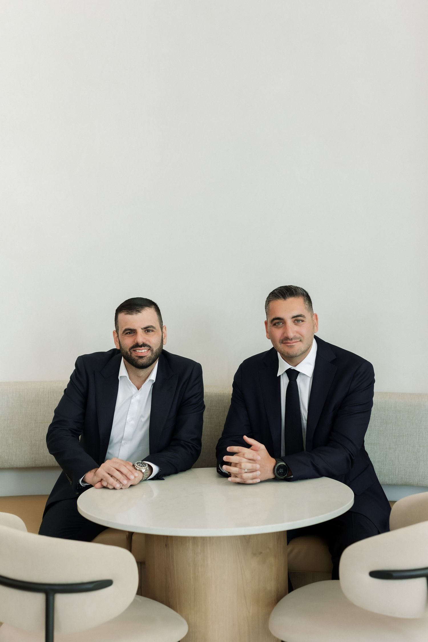 Two professional men in dark suits sit at a round table in a modern office setting, smiling at the camera.