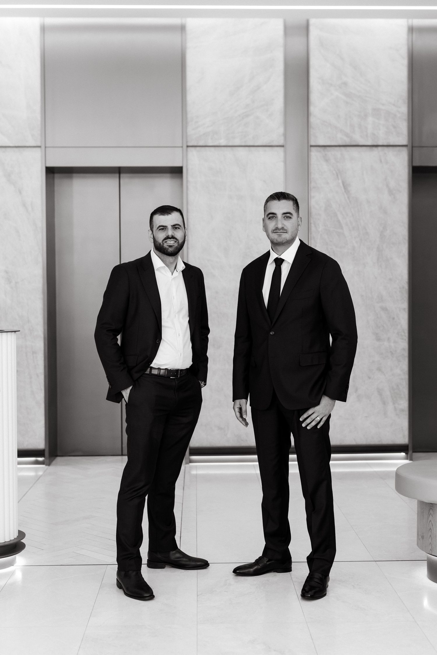 Two professional men in black suits stand confidently in a modern marble lobby for a formal business portrait.