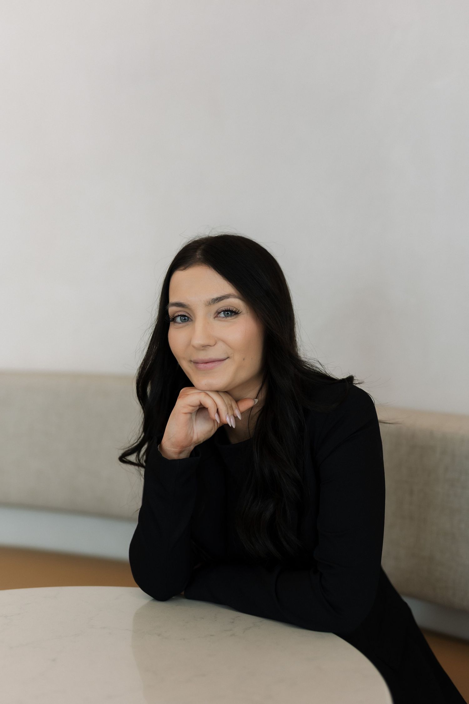 Young woman with dark hair in black top sitting at white table in modern minimalist space.
