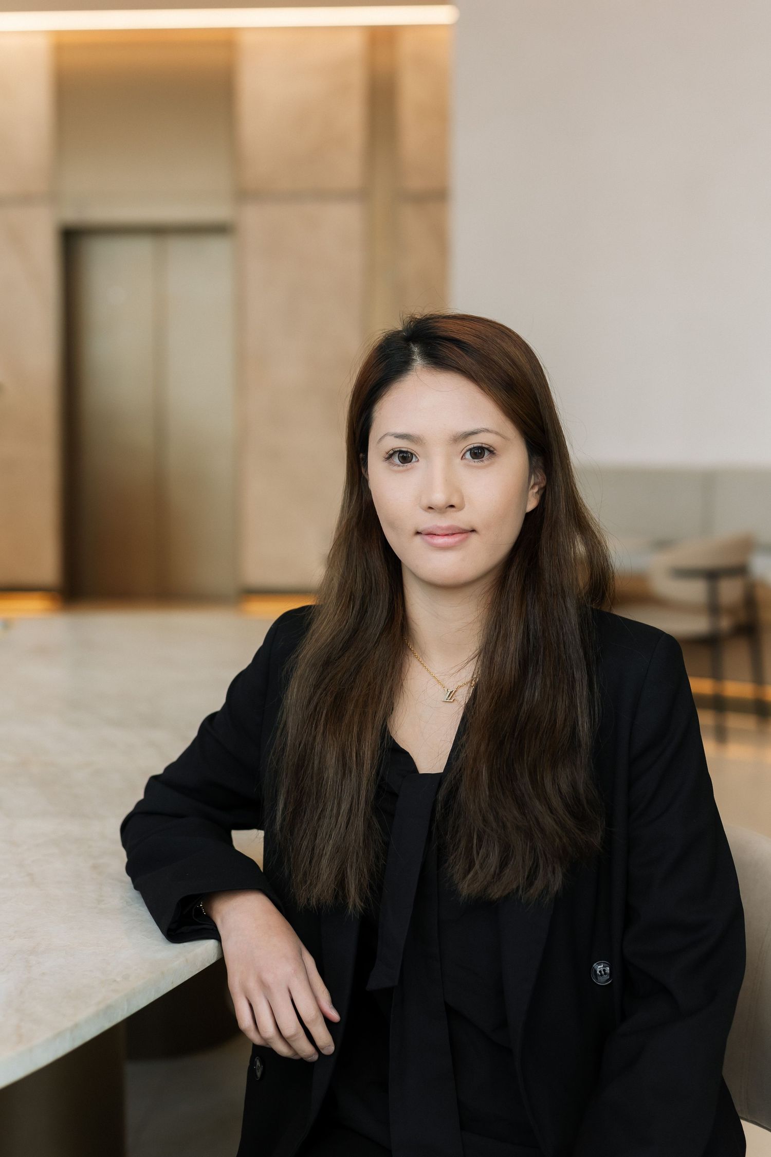 Young professional woman in black blazer leaning on marble counter in modern lobby setting.