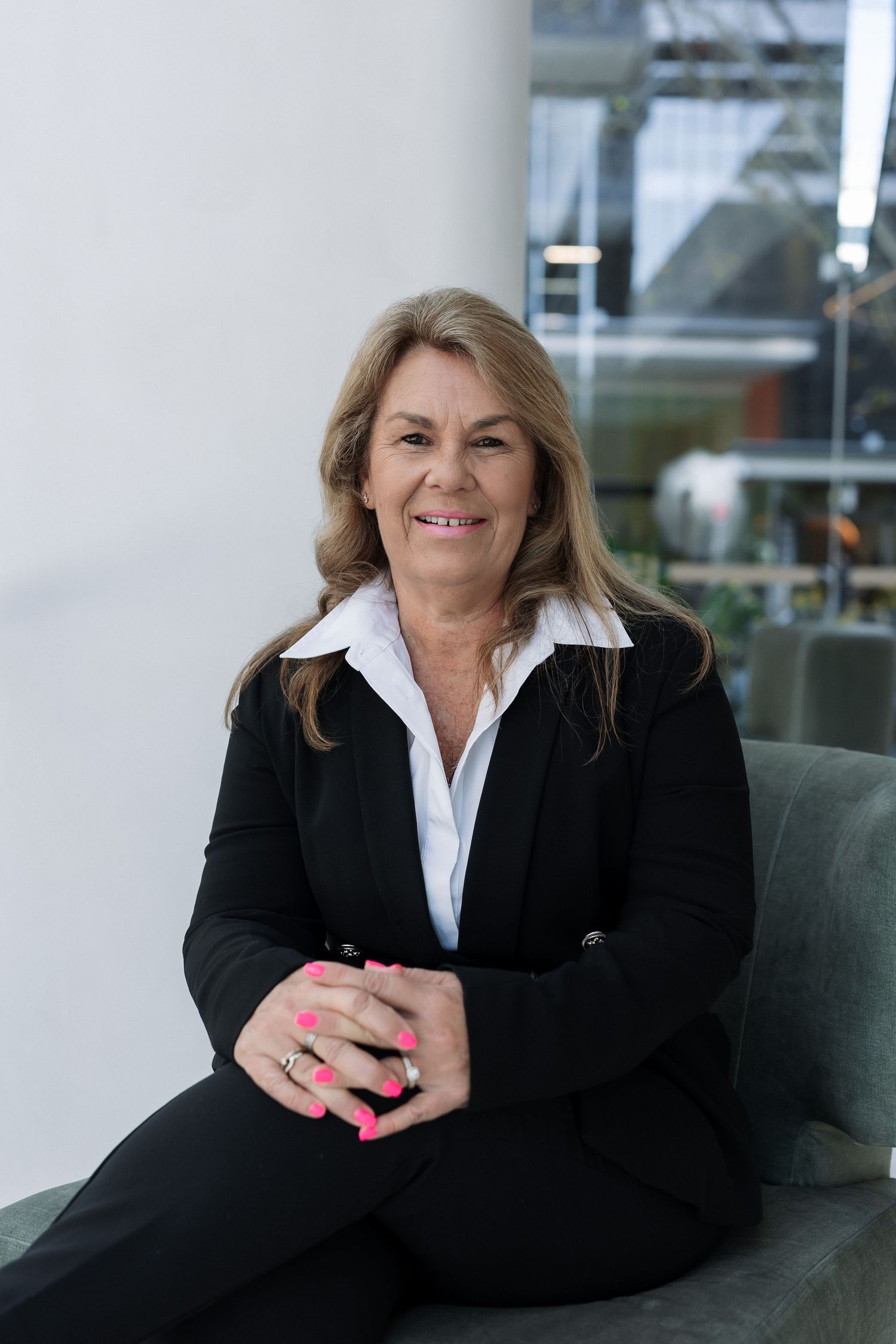Professional businesswoman in black blazer sitting confidently with clasped hands, smiling in modern office setting.