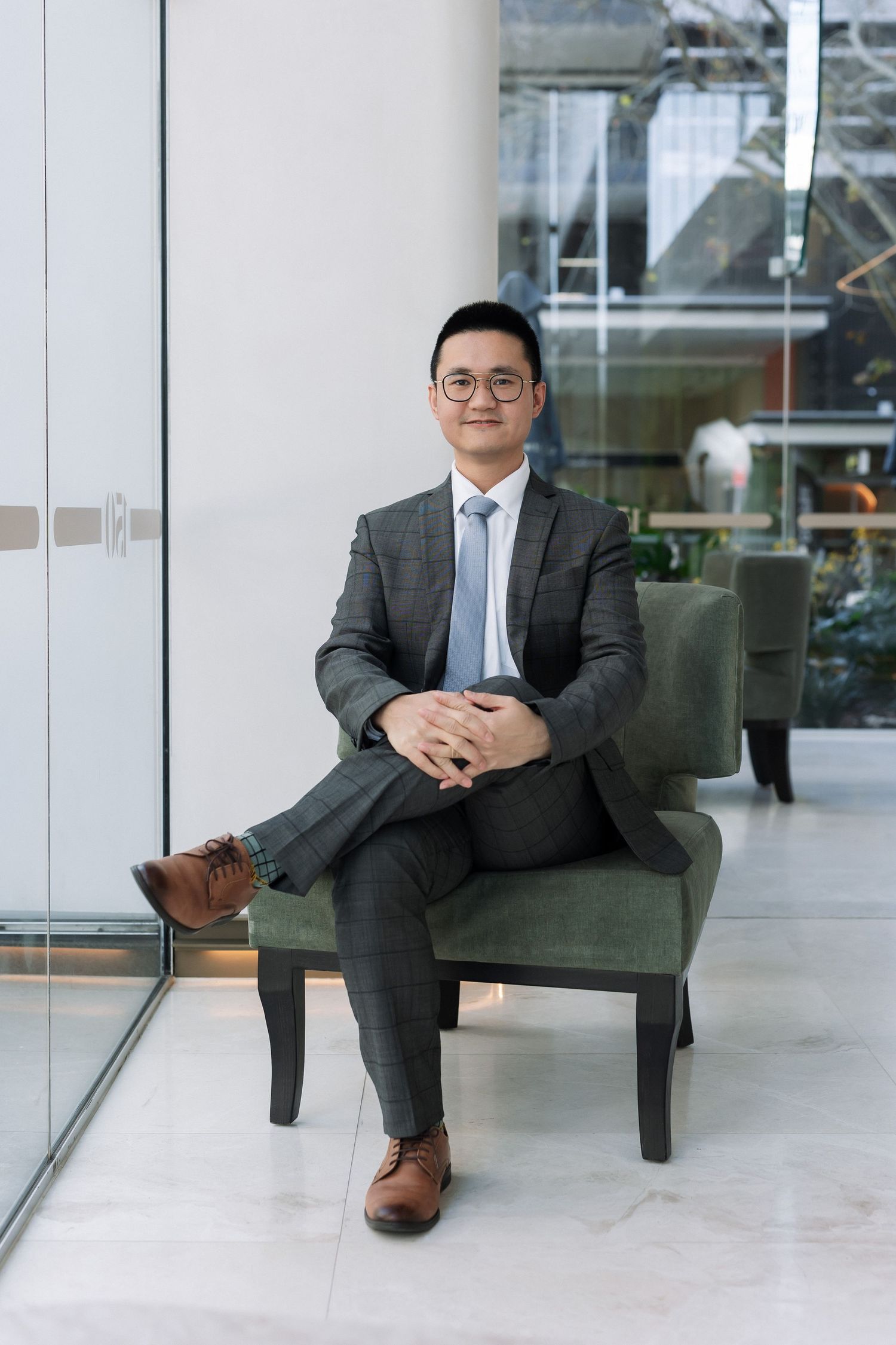 Professional Asian man in gray suit sitting confidently in green armchair in modern office lobby.