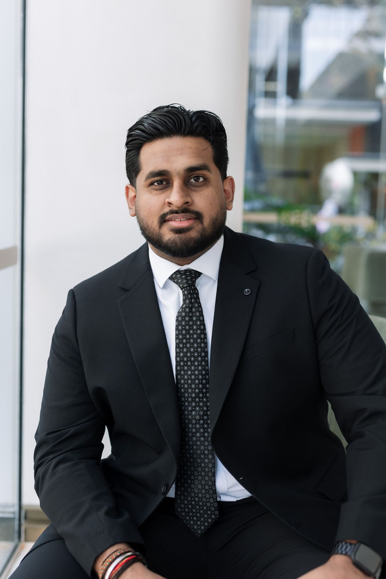 Young businessman in black suit and dotted tie seated in modern office lobby, professional portrait.
