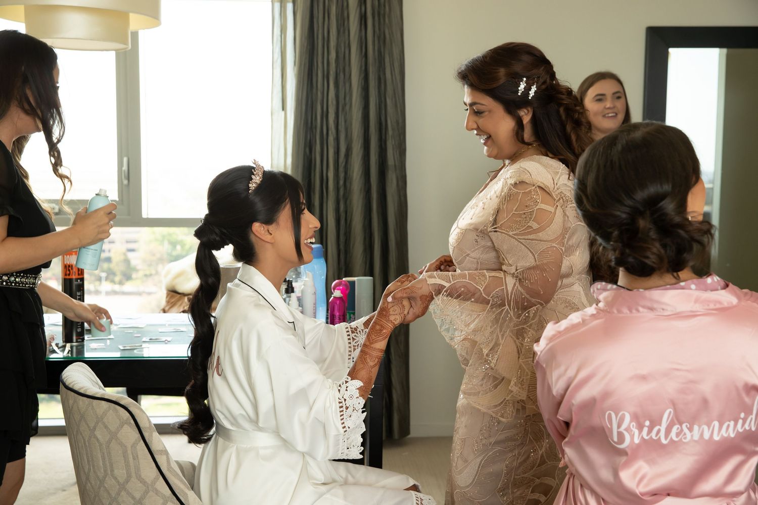 Bride in lace gown laughing with bridesmaids in satin robes during wedding morning preparations.