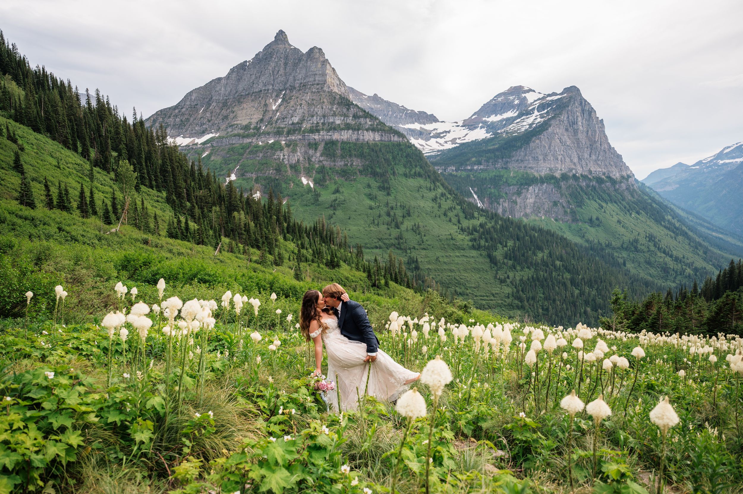 Couple sharing a romantic kiss in a wildflower meadow with stunning mountain peaks in Glacier National Park.