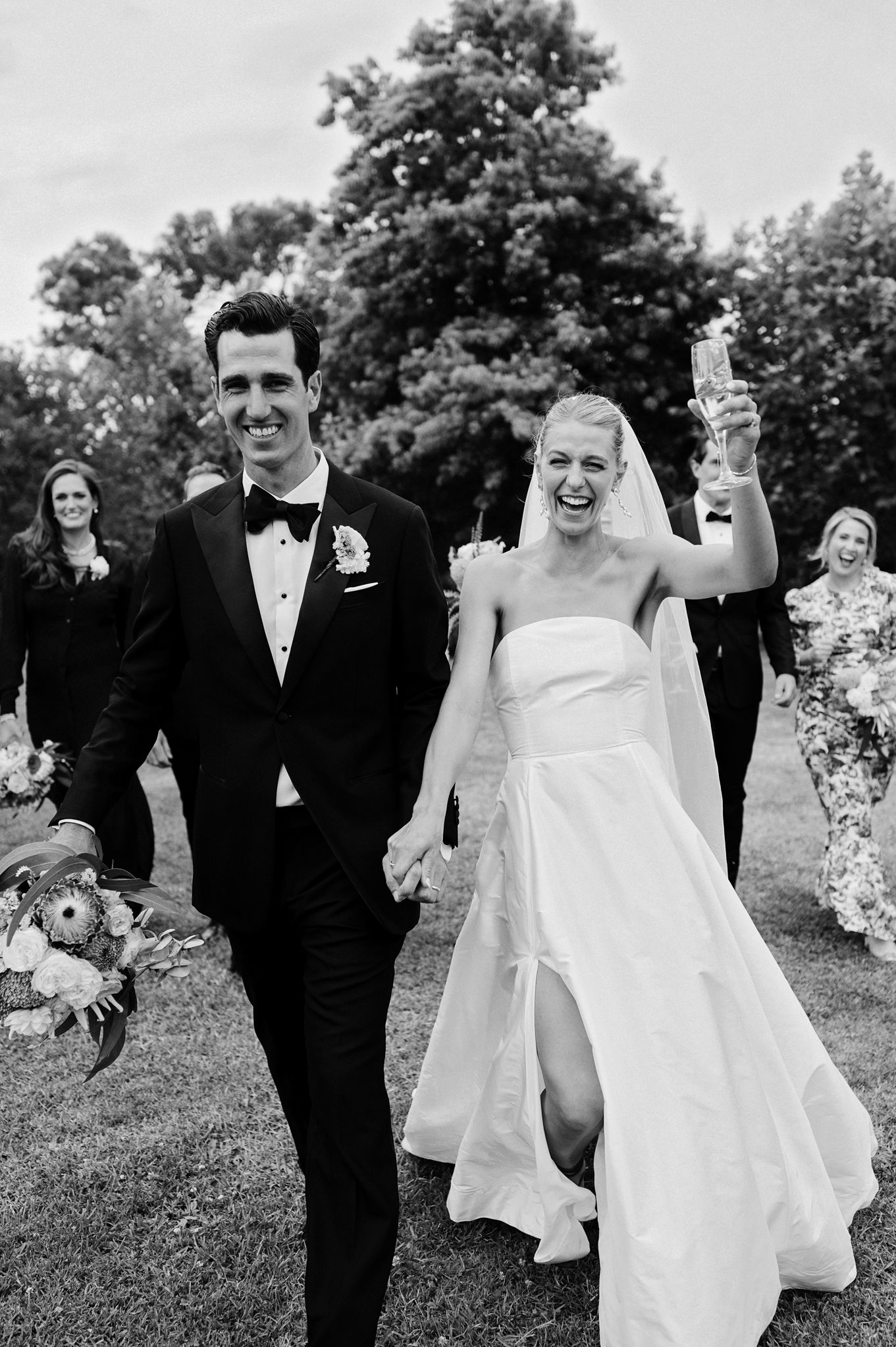 Joyful newlyweds walk hand in hand after ceremony, bride raising champagne glass, black and white photo.