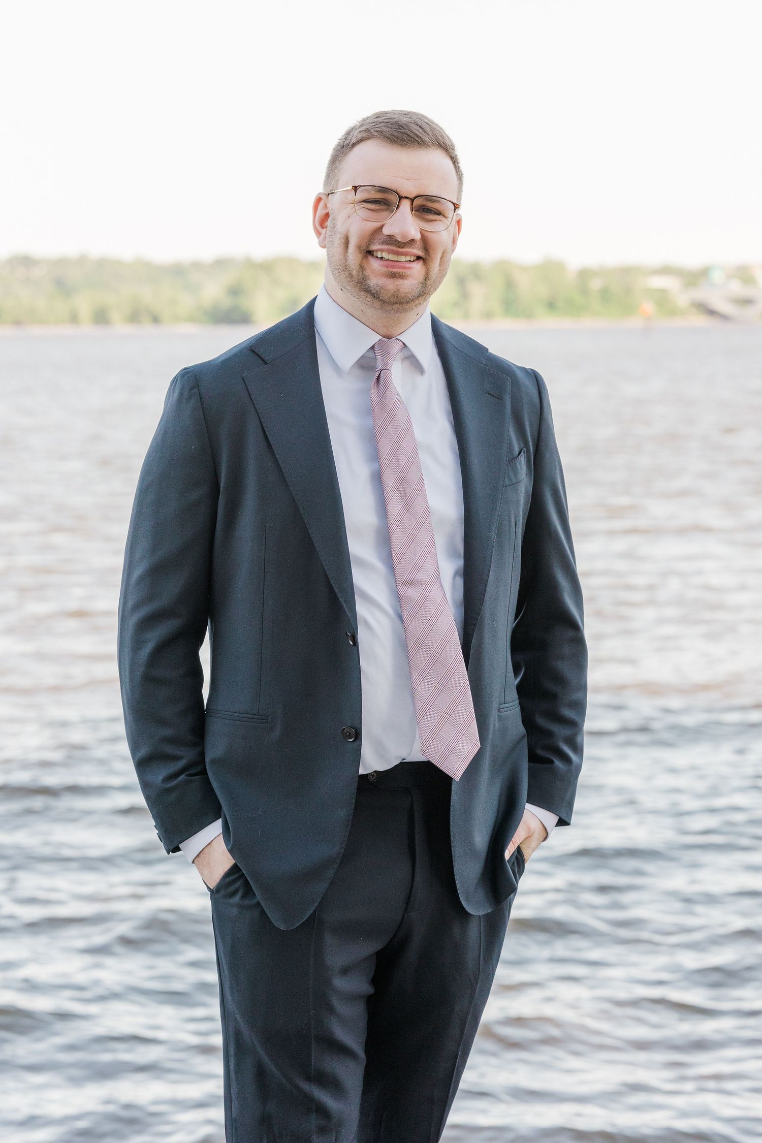 Smiling businessman in dark suit and pink tie standing outdoors near water with hands in pockets.