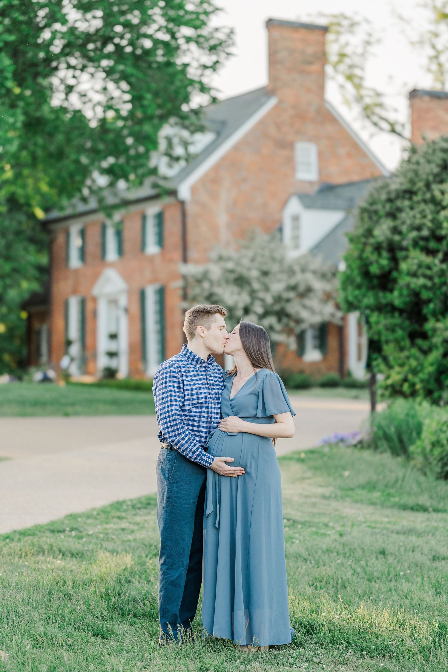 A couple shares a tender kiss outdoors, the woman in a blue maternity gown cradling her bump near a brick estate.