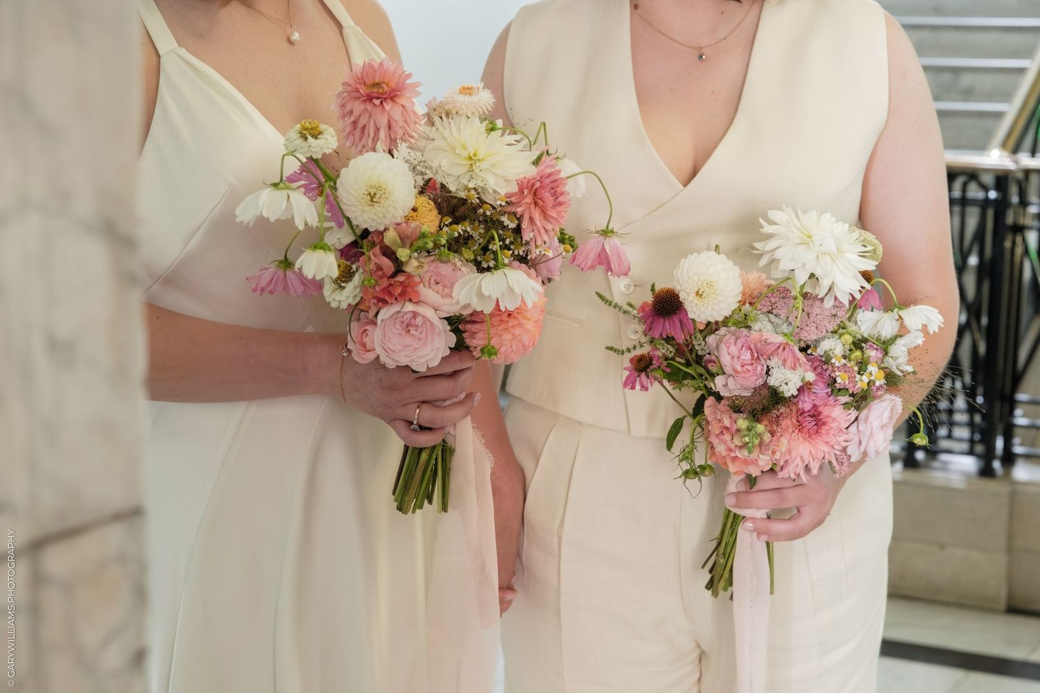 Two bridesmaids in cream outfits holding pink and white floral bouquets with dahlias and wildflowers.