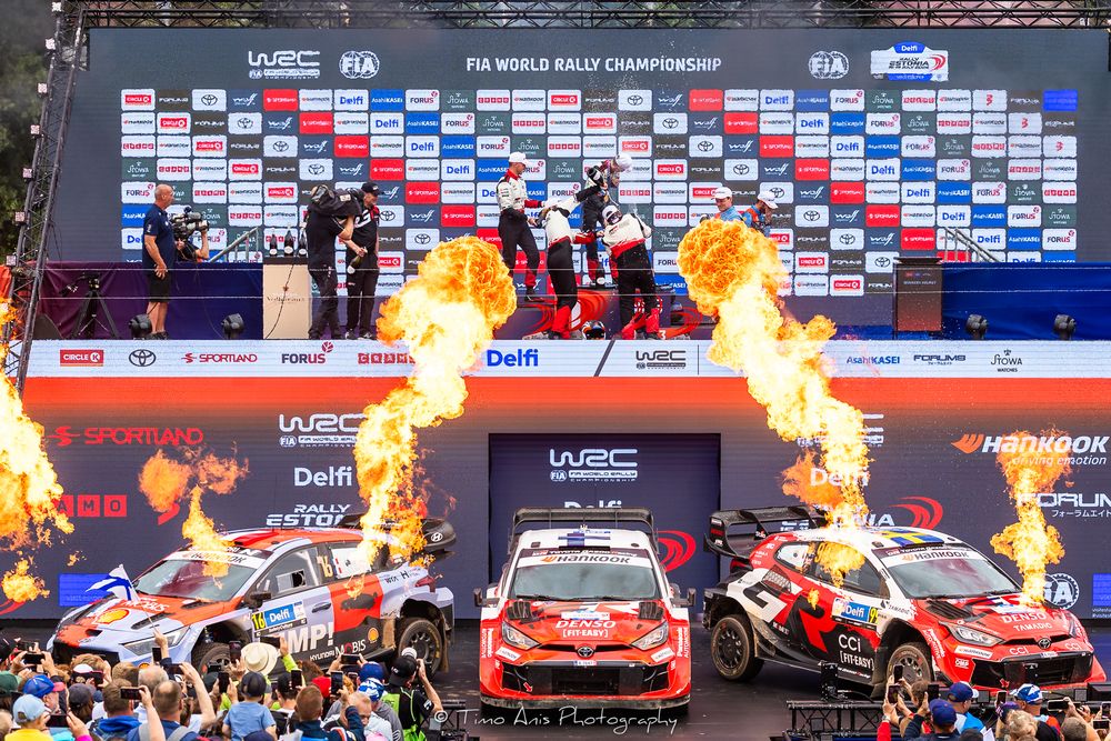 FIA World Rally Championship podium celebration with fire displays, rally cars, and trophy presentation at an outdoor racing event.