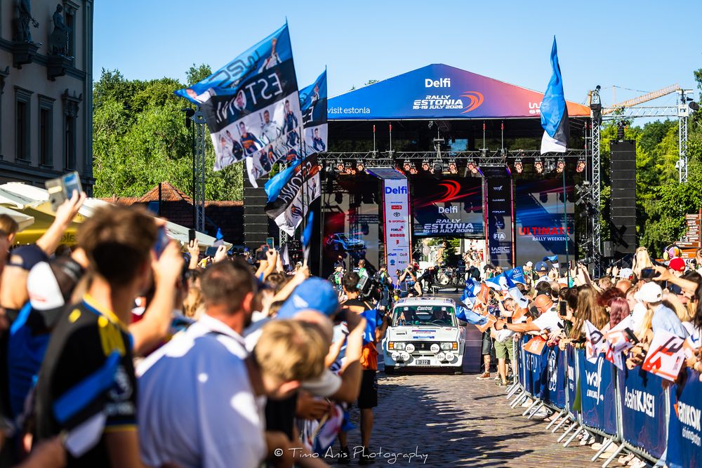 Rally car drives through cheering crowd at outdoor motorsport event with colorful flags and sponsor banners on sunny day.