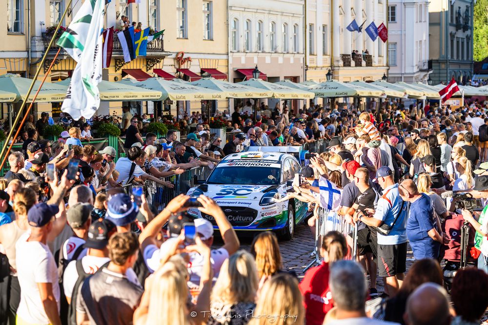 Rally car navigates through a crowded town square lined with spectators during a WRC rally event.