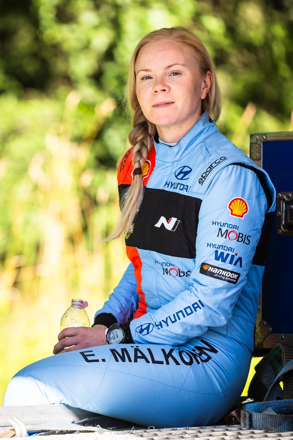 Rally driver E. Malkon poses in a blue Hyundai Mobis racing suit with Shell and Hankook sponsors outdoors.