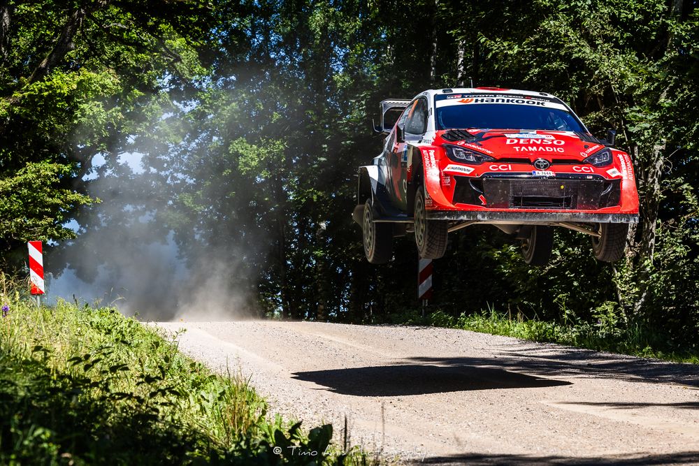 Red rally car catches air mid-jump on a forest stage road during a competitive rally racing event.