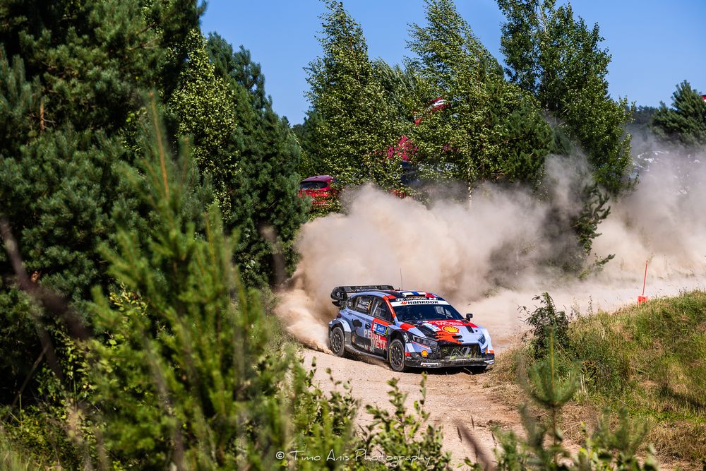 Rally car racing on a dusty gravel forest road, kicking up clouds of dust during a WRC rally stage competition.