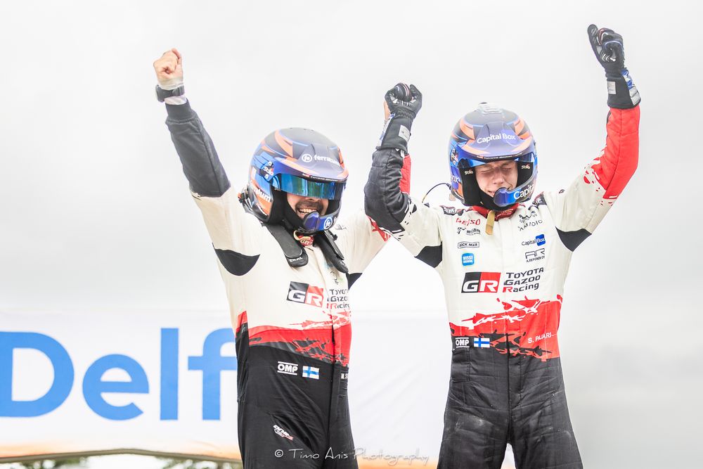 Two rally drivers celebrate victory with raised arms on podium at Delft motorsport event wearing helmets and racing suits.