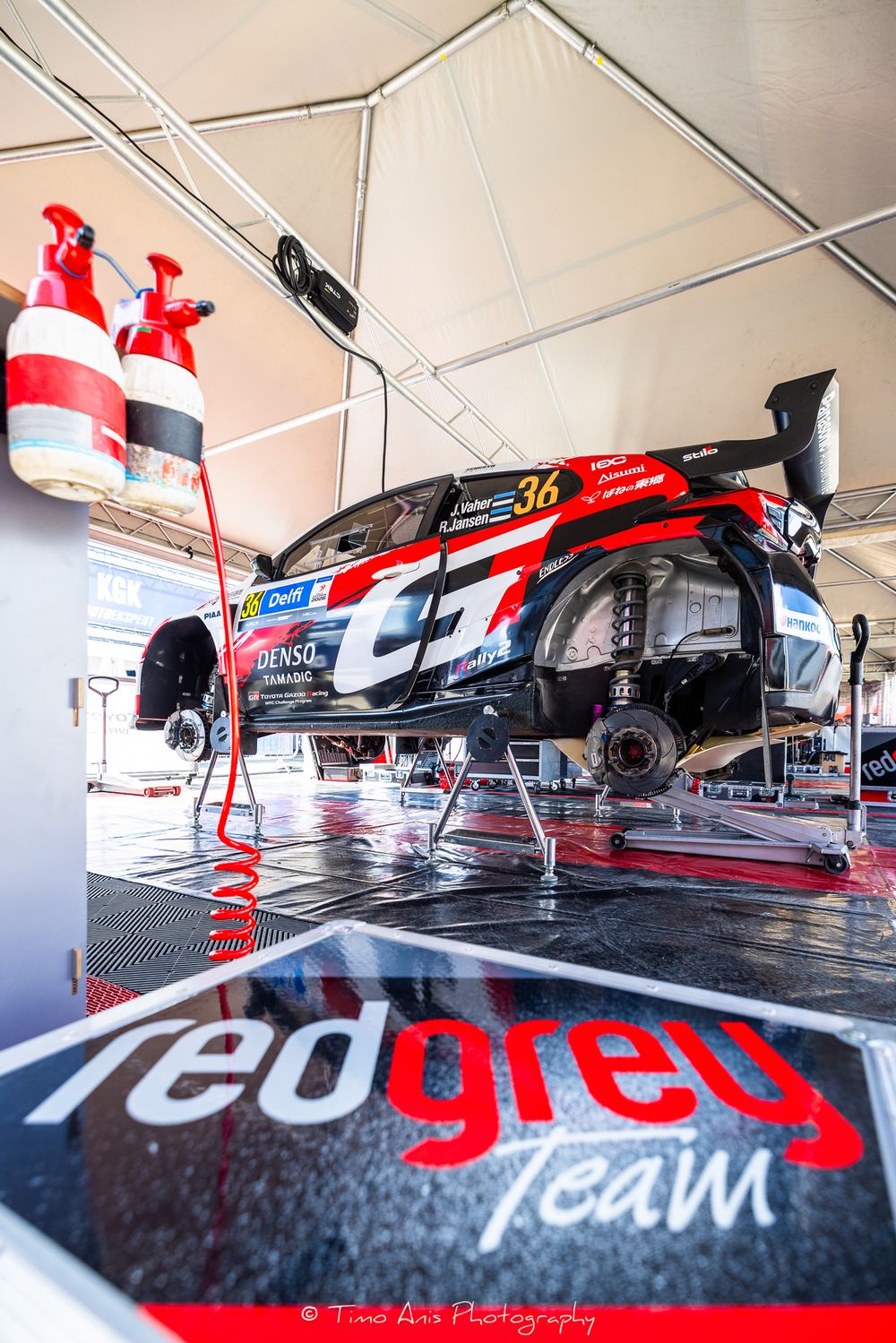 Red Grey Team rally car #36 raised on jacks inside a service tent with fire extinguishers visible in the foreground.