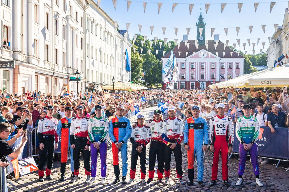 Racing drivers lineup at a Formula E event in a historic European town square surrounded by crowds.