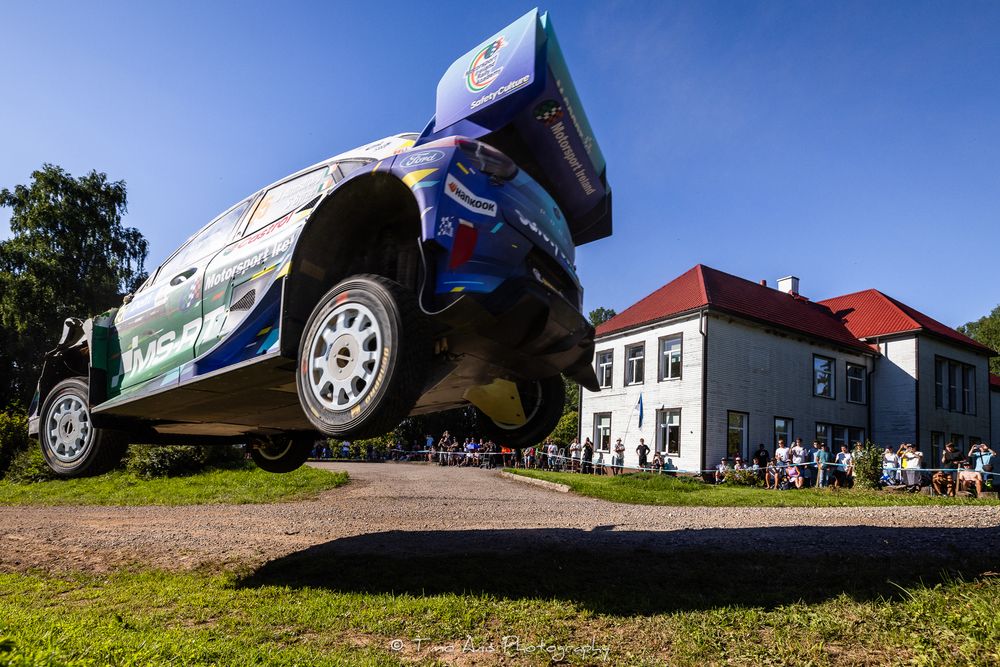 Rally car catches massive air during a jump at a WRC stage race event with spectators watching nearby.