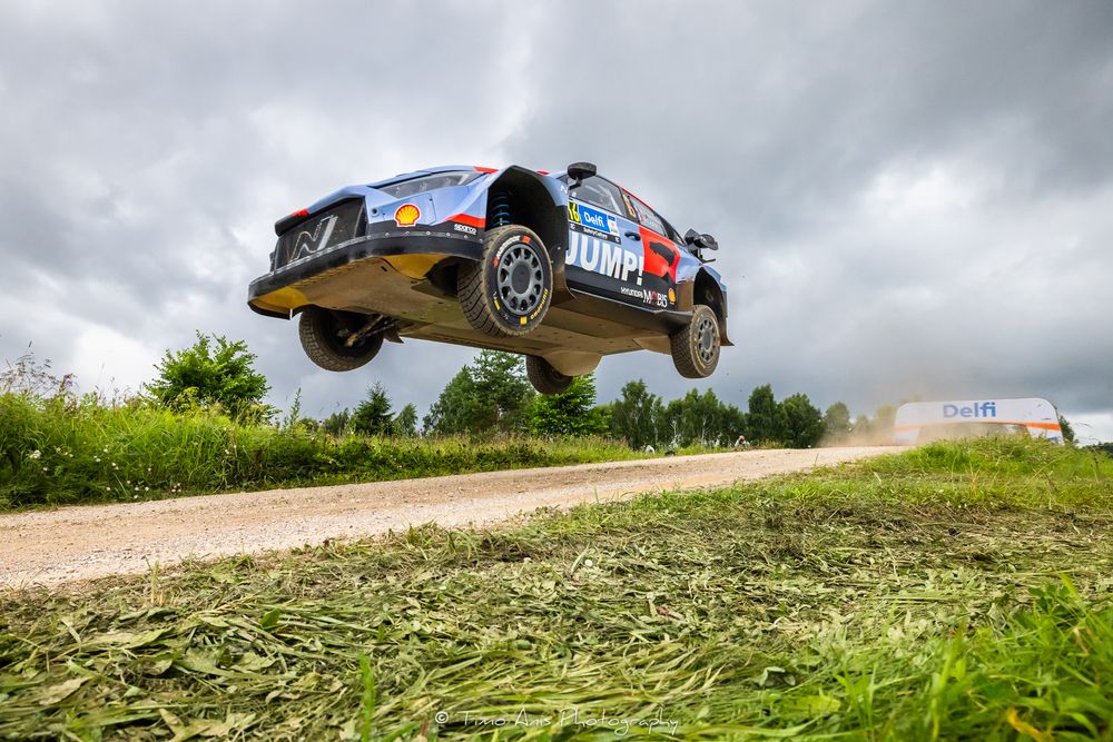 Rally car soaring through the air on a dirt road during a motorsport competition under dramatic cloudy skies.