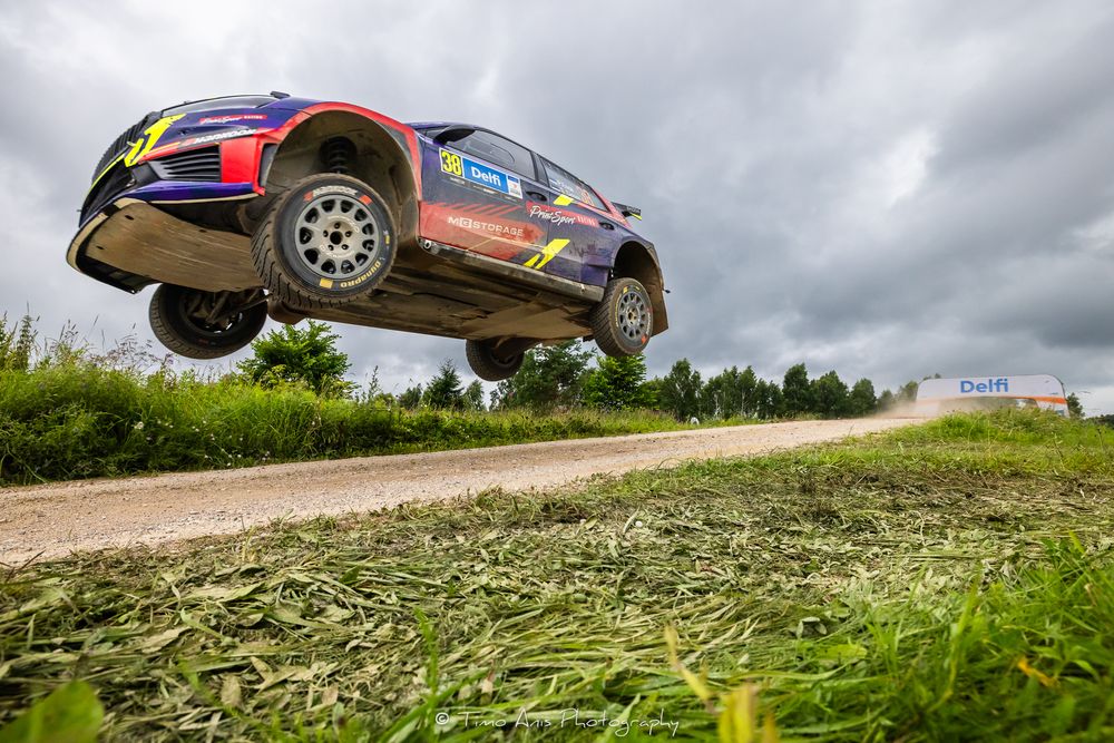 A rally car catches massive air on a gravel road during a WRC rally stage under cloudy skies.