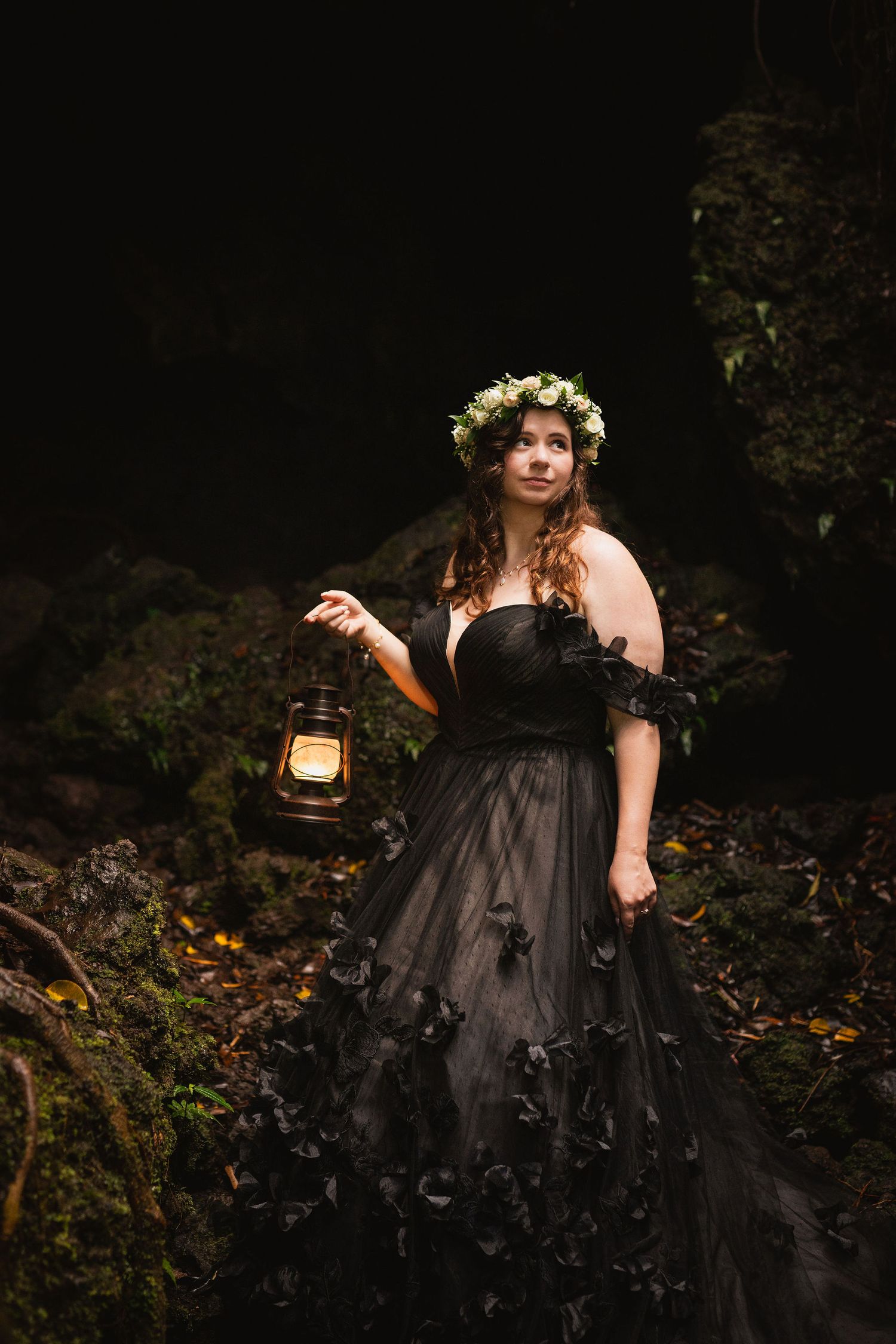 Woman in black gown with floral crown holding a lantern in a dark forest.