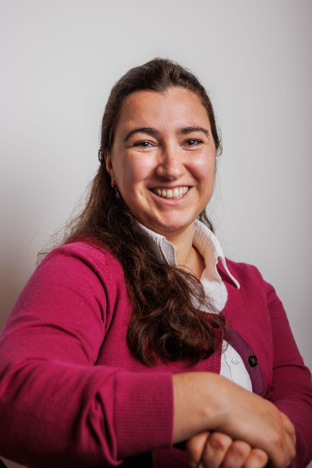 Smiling young woman with dark hair wearing a pink cardigan, posing with arms crossed in a professional portrait.