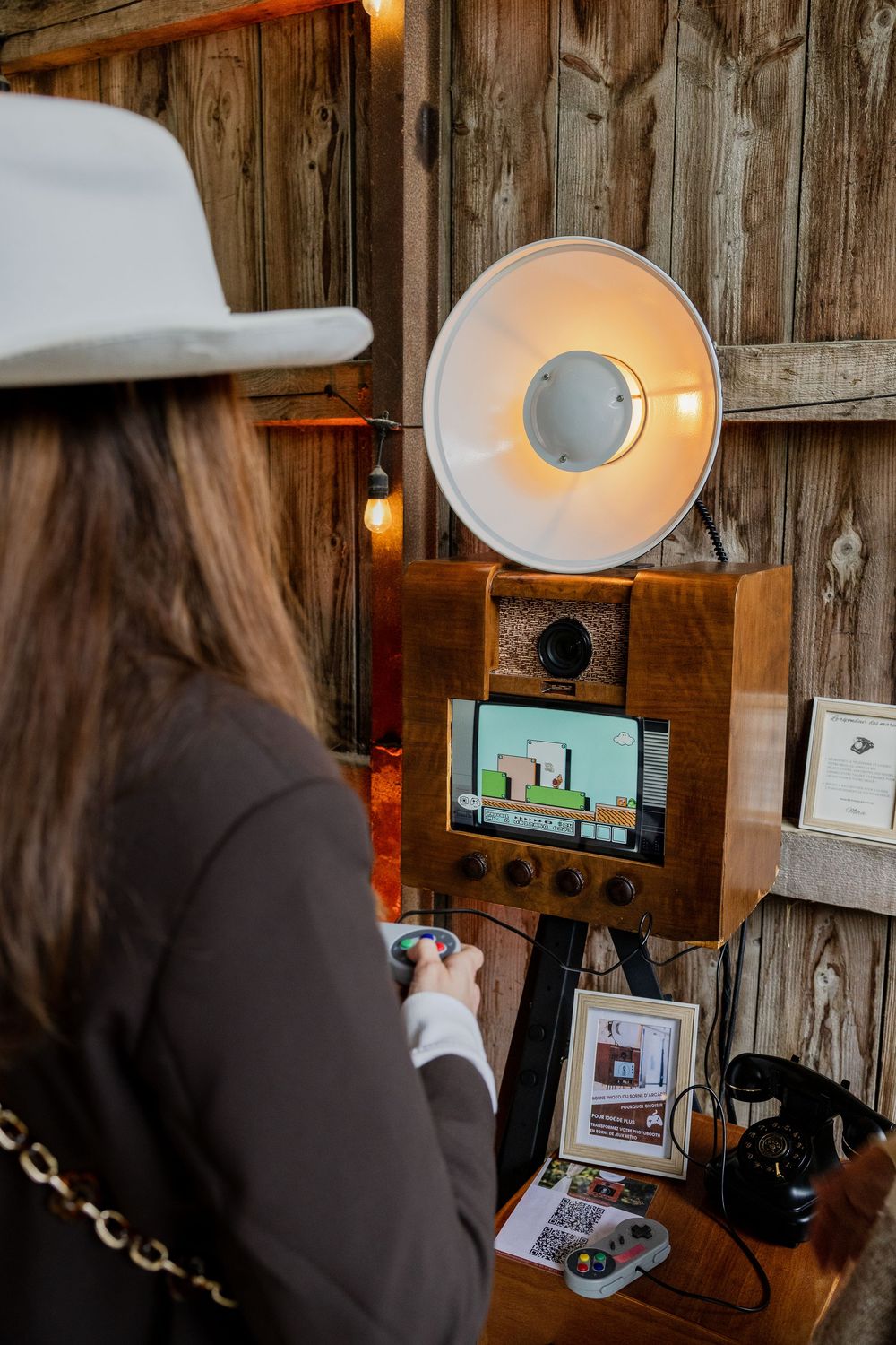 Woman in white hat using a vintage wooden photo booth with large flash lamp at a rustic event.