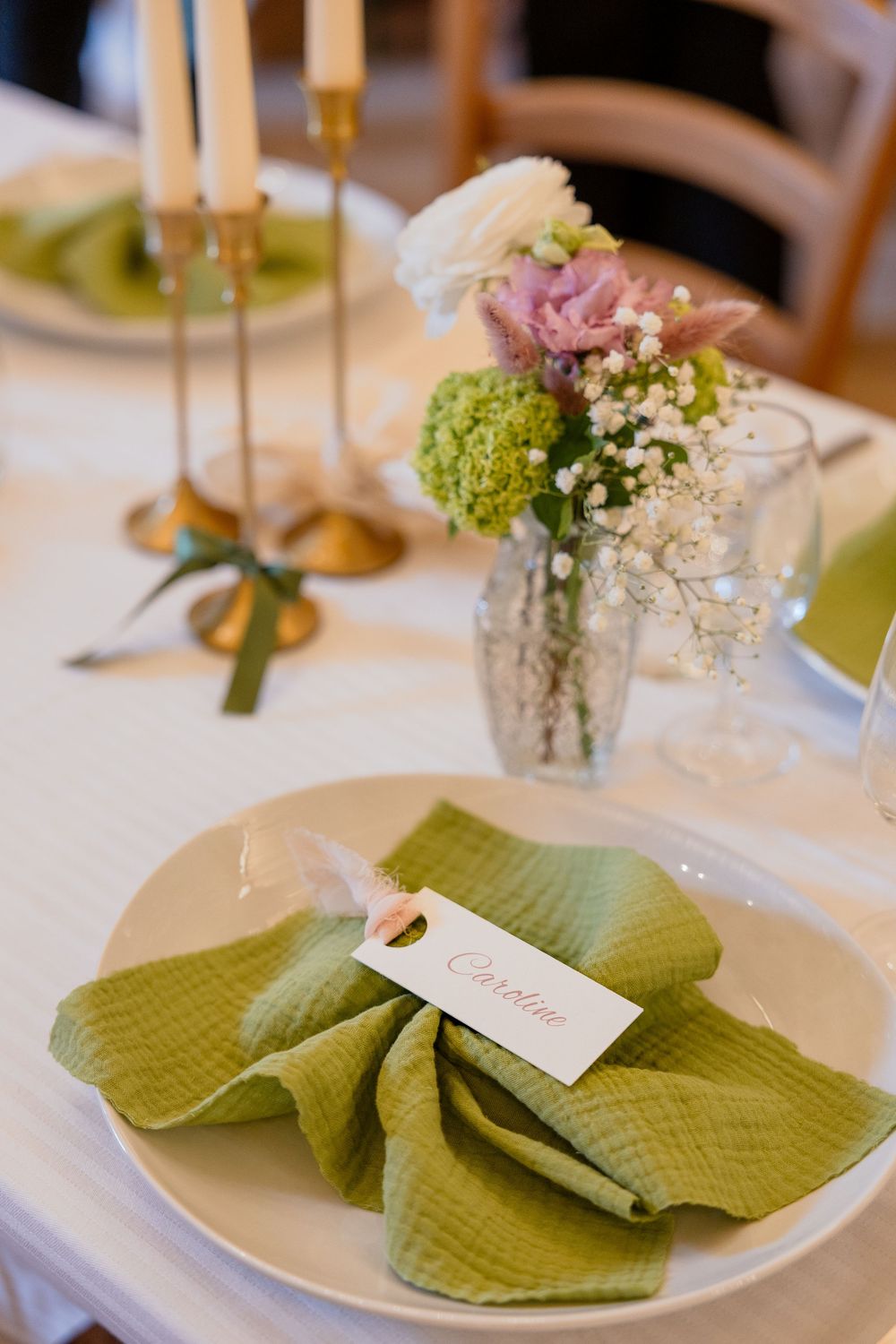 Elegant wedding table setting with green linen napkin, name card reading Caroline, floral centerpiece, and gold candlesticks.