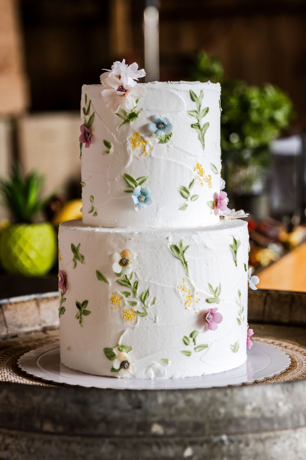Two-tier white wedding cake with hand-painted floral details in pink, blue, and yellow on a rustic barrel stand.
