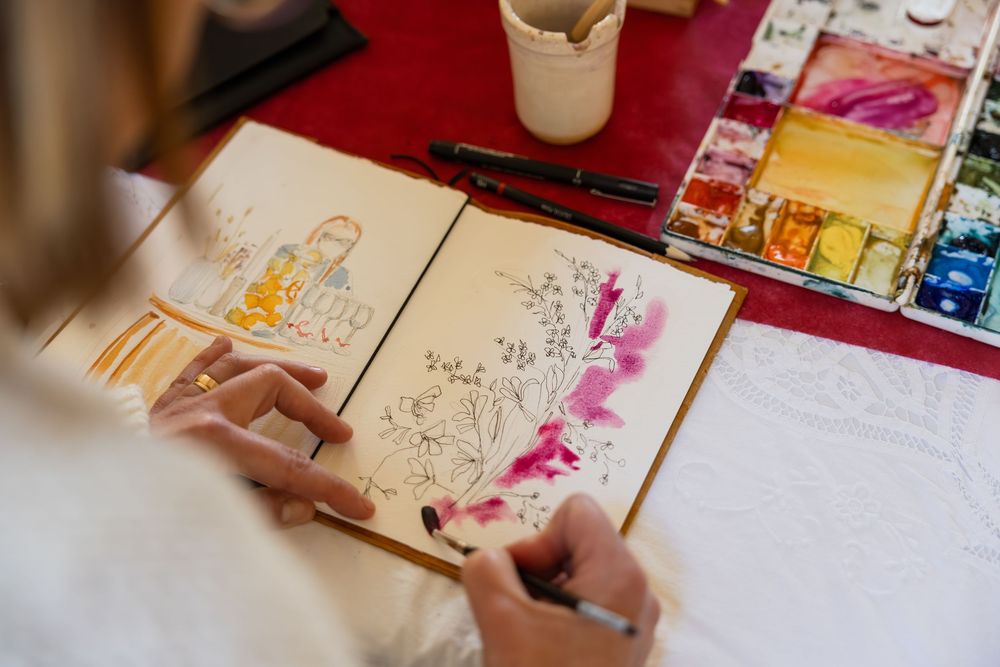 Artist painting floral watercolor sketches in an open sketchbook with colorful paints and brushes on a red table.