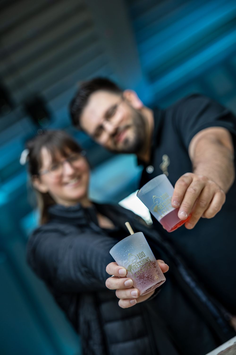 Two smiling people in black clothes holding colorful drinks toward the camera outdoors.