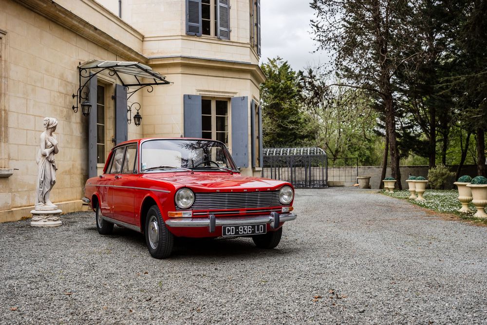 Vintage red classic car parked on gravel driveway outside elegant French chateau with stone statue.
