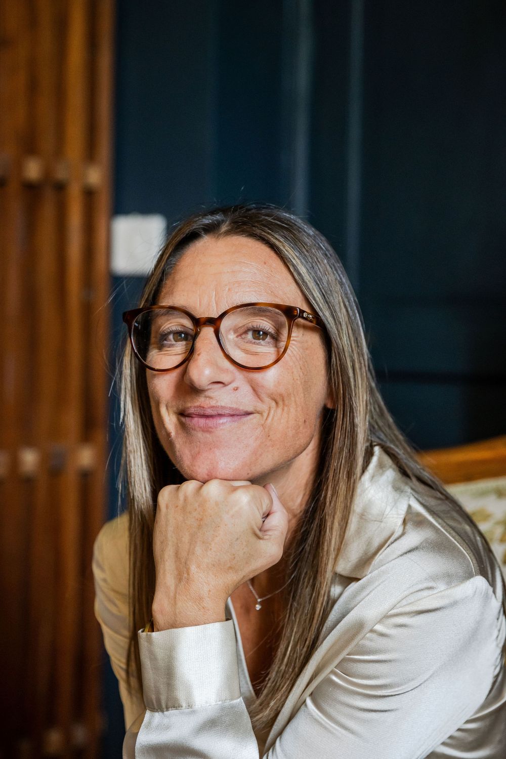Smiling middle-aged woman with long hair and tortoiseshell glasses, resting chin on hand, wearing a satin blazer.