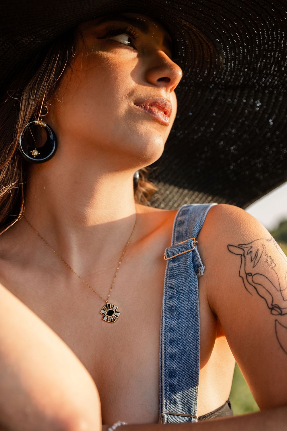 Woman in black sun hat and denim overalls wearing celestial sun necklace and earrings with arm tattoo in golden light.