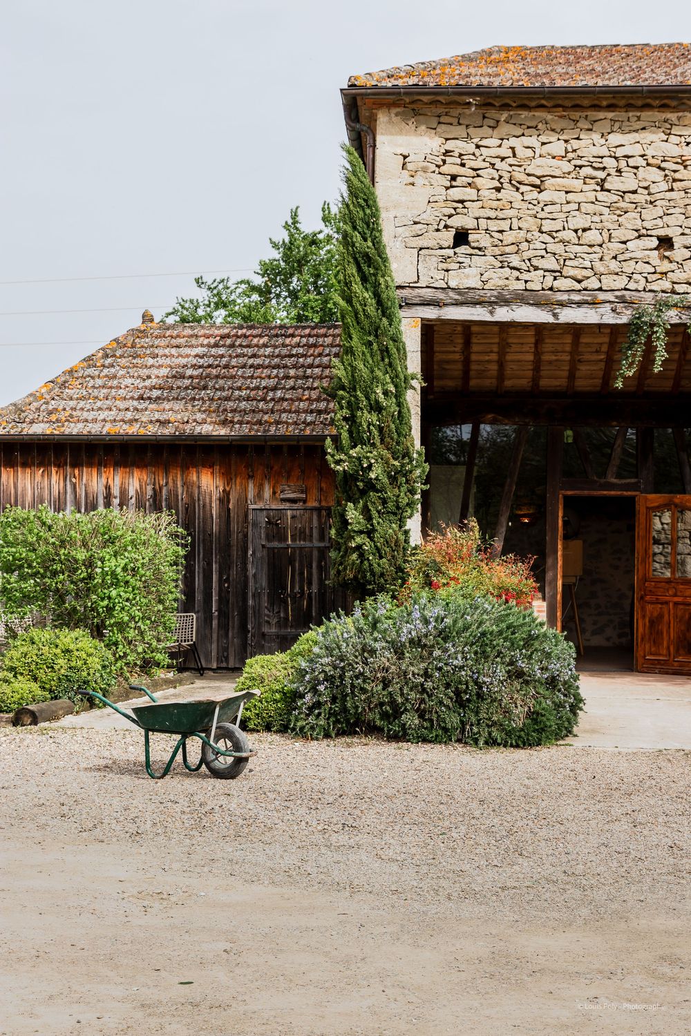 Rustic stone farmhouse with wooden barn, wheelbarrow, and lush garden shrubs on a gravel courtyard.