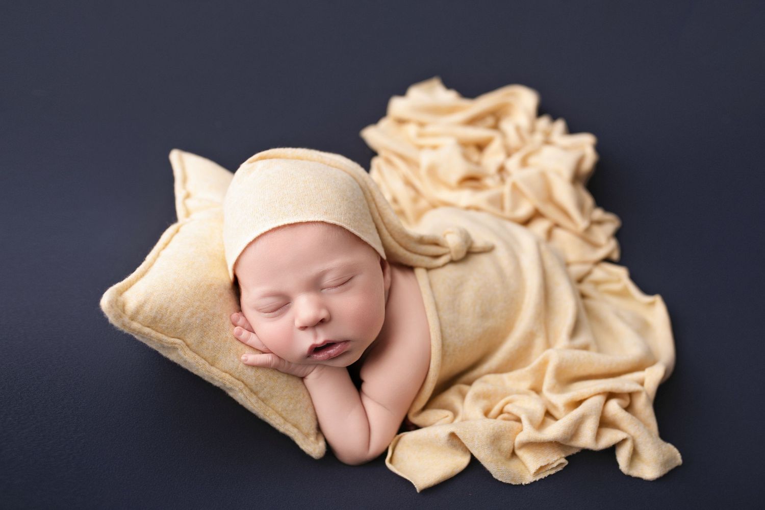Newborn baby sleeping peacefully on a cream pillow, wrapped in soft yellow fabric with a matching bonnet.